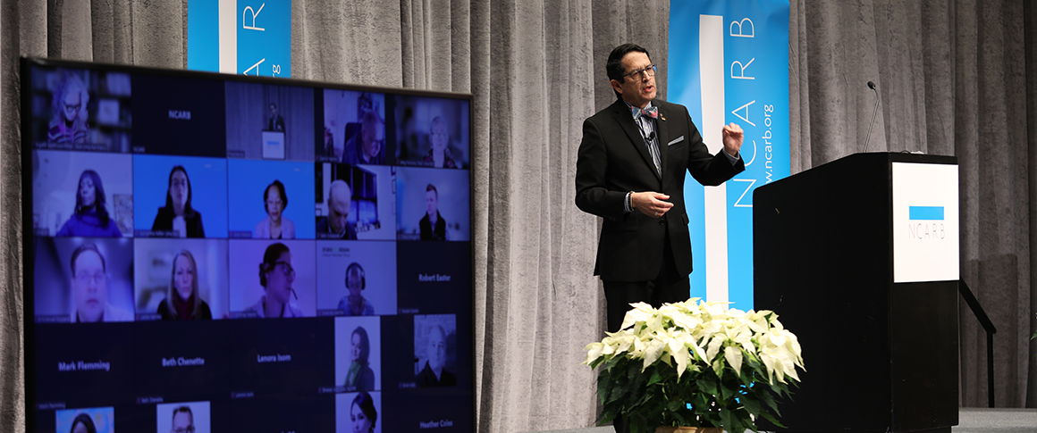 Alfred Vidaurri presents from a podium to virtual and in person attendees.