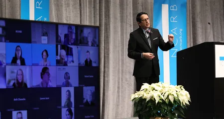Alfred Vidaurri presents from a podium to virtual and in person attendees.