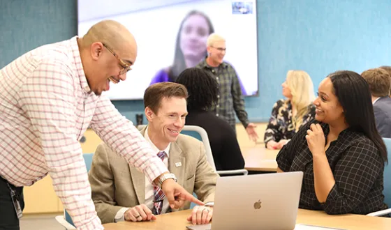 Three NCARB staff members collaborate around a laptop during a hybrid meeting. More staff members are in the background.