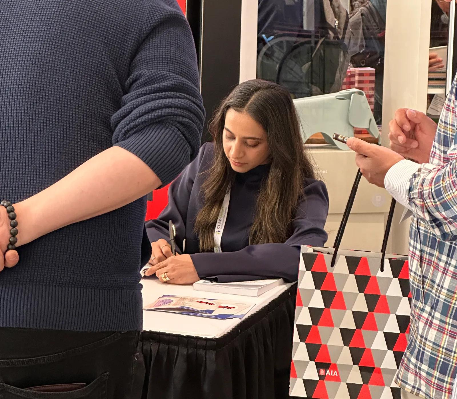 Apurva sits at a table while signing a copy of the book she co-authored, City Shapers.